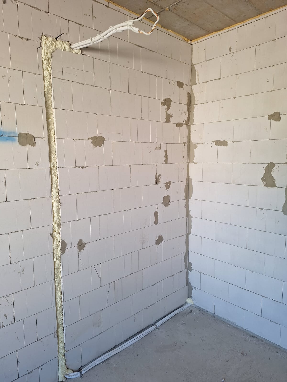 Wewnętrzna ściana z betonu komórkowego w trakcie prac instalacyjnych, widoczne bruzdy wypełnione pianką montażową, rury instalacyjne prowadzone wzdłuż ścian i pod sufitem, surowy stan wykończenia.
