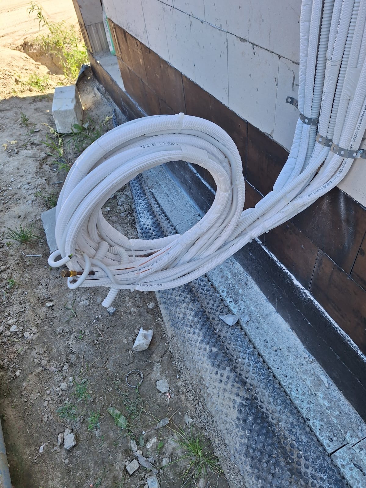 Zwój białych rur klimatyzacyjnych leżący na ziemi obok fundamentu domu w trakcie instalacji.