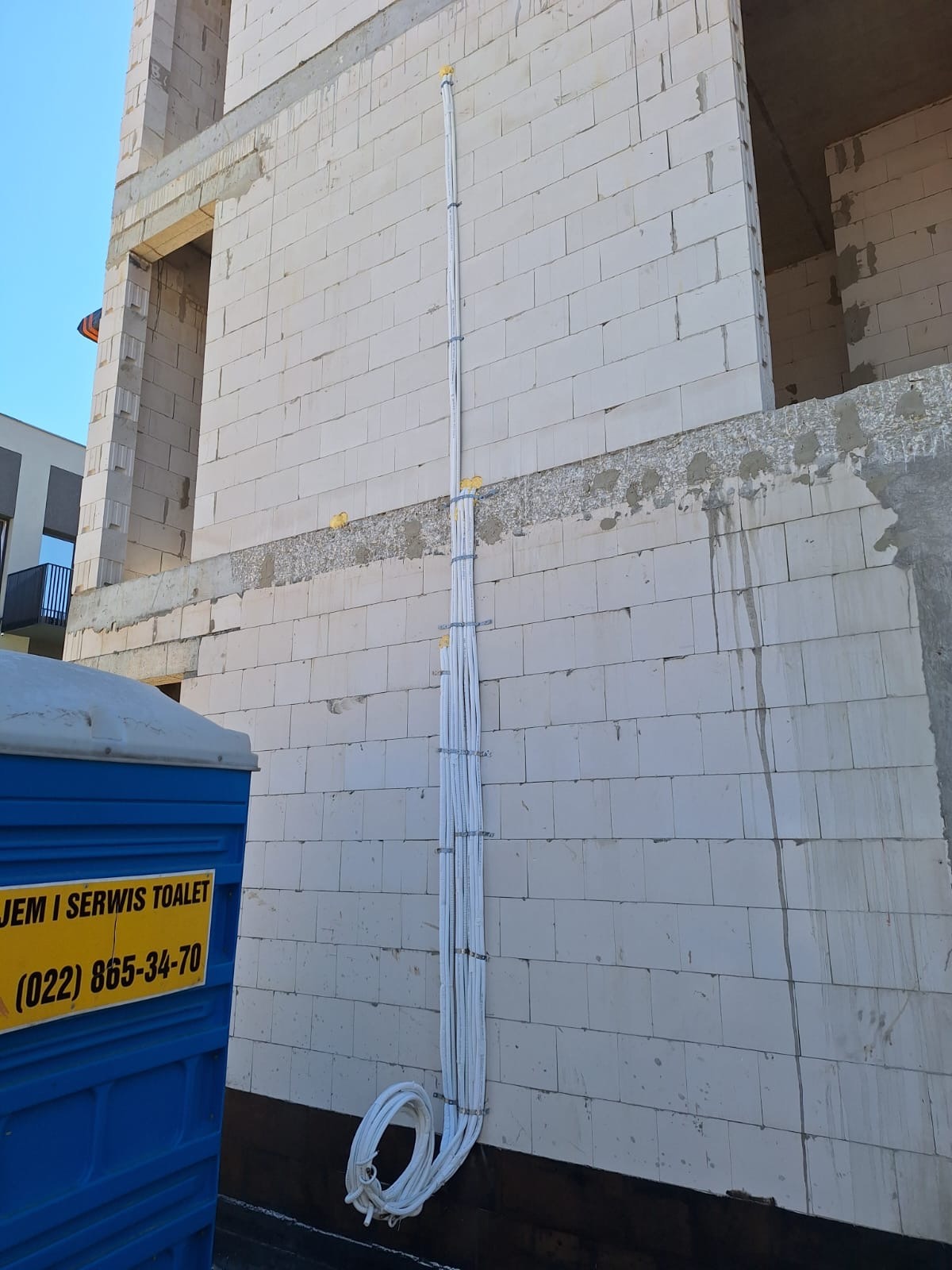 Białe rury wentylacyjne przymocowane do ściany budynku w trakcie budowy, obok niebieska toaleta przenośna.