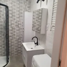 Nowoczesna łazienka z heksagonalnymi płytkami w odcieniach szarości, czarną armaturą, białą umywalką z szafką, pralką i grzejnikiem drabinkowym.