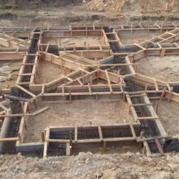 Przygotowanie szalunku i zbrojenia pod fundament budynku, widoczne drewniane deski i stalowe pręty, wykop ziemny.