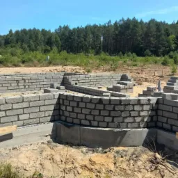 Budowa fundamentów z bloczków betonowych, widoczny zarys ścian i fragment z zaokrąglonym narożnikiem, w tle las i częściowo widoczny budynek z czerwonym dachem, na placu budowy osoba z taczką.