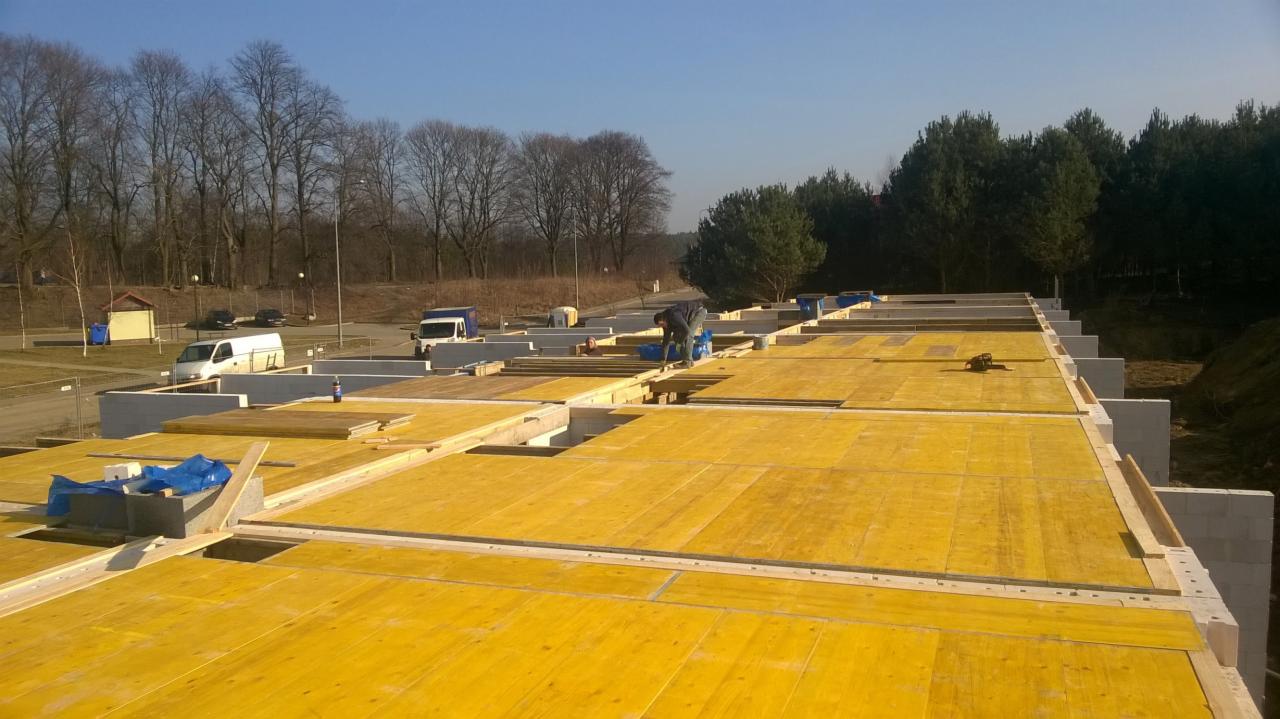 Budowa domu z bloczków betonu komórkowego, widok z góry na przygotowany strop z żółtymi płytami szalunkowymi, na budowie pracuje robotnik, w tle drzewa i zaparkowane samochody.