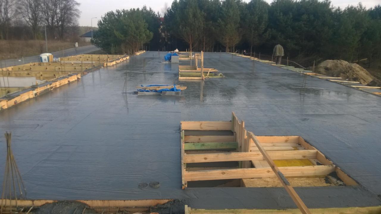 Wylany beton na stropie budynku w trakcie budowy, widoczne drewniane szalunki, zbrojenia oraz osoba w oddali.