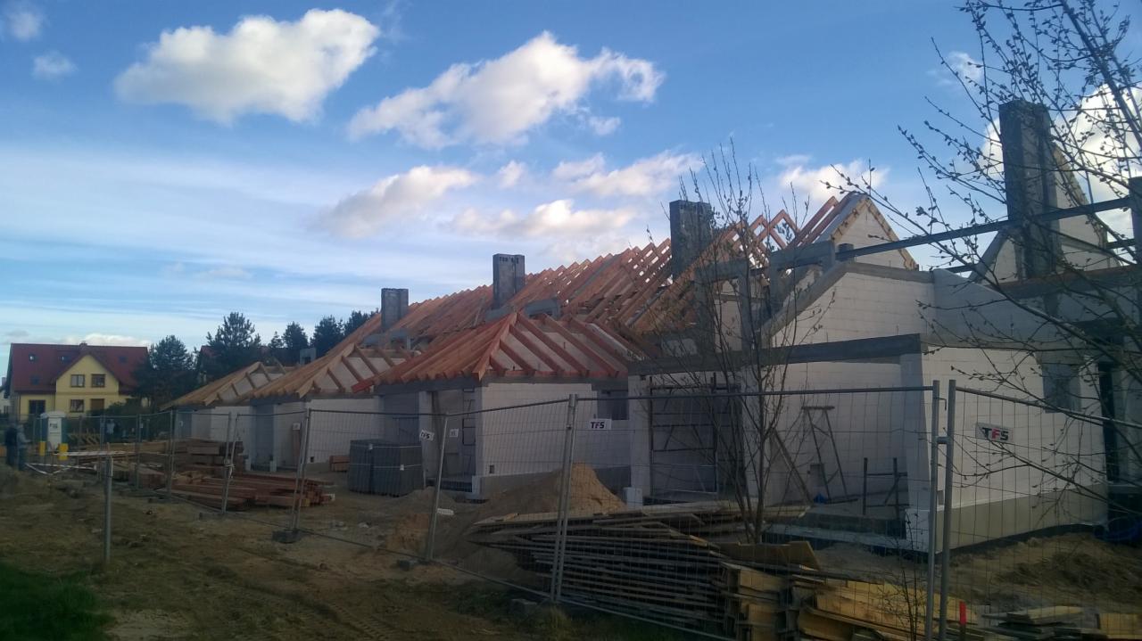 Wznoszenie szeregu budynków jednorodzinnych z czerwonymi dachówkami, widoczne drewniane konstrukcje dachowe, kominy, ściany z białego bloczka oraz materiały budowlane składowane na placu budowy...