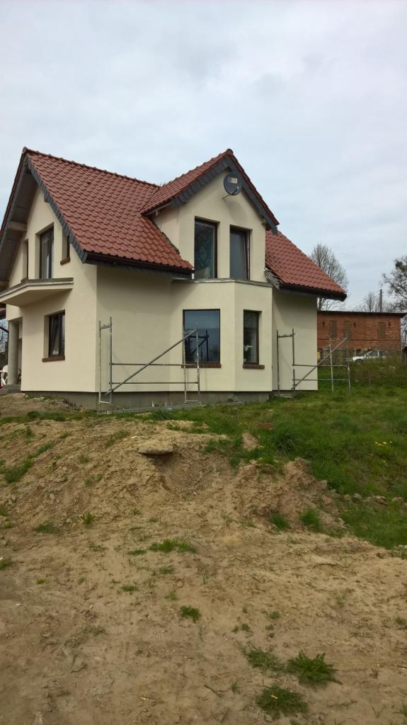 Nowo wybudowany, otynkowany dom z czerwoną dachówką, widoczny z poziomu ziemi, z rusztowaniem przy ścianie i stertą ziemi na pierwszym planie.
