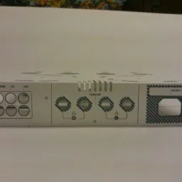 Metalowa obudowa urządzenia elektronicznego z precyzyjnymi nadrukami technicznymi, widoczne otwory wentylacyjne i panel z opisami funkcji.