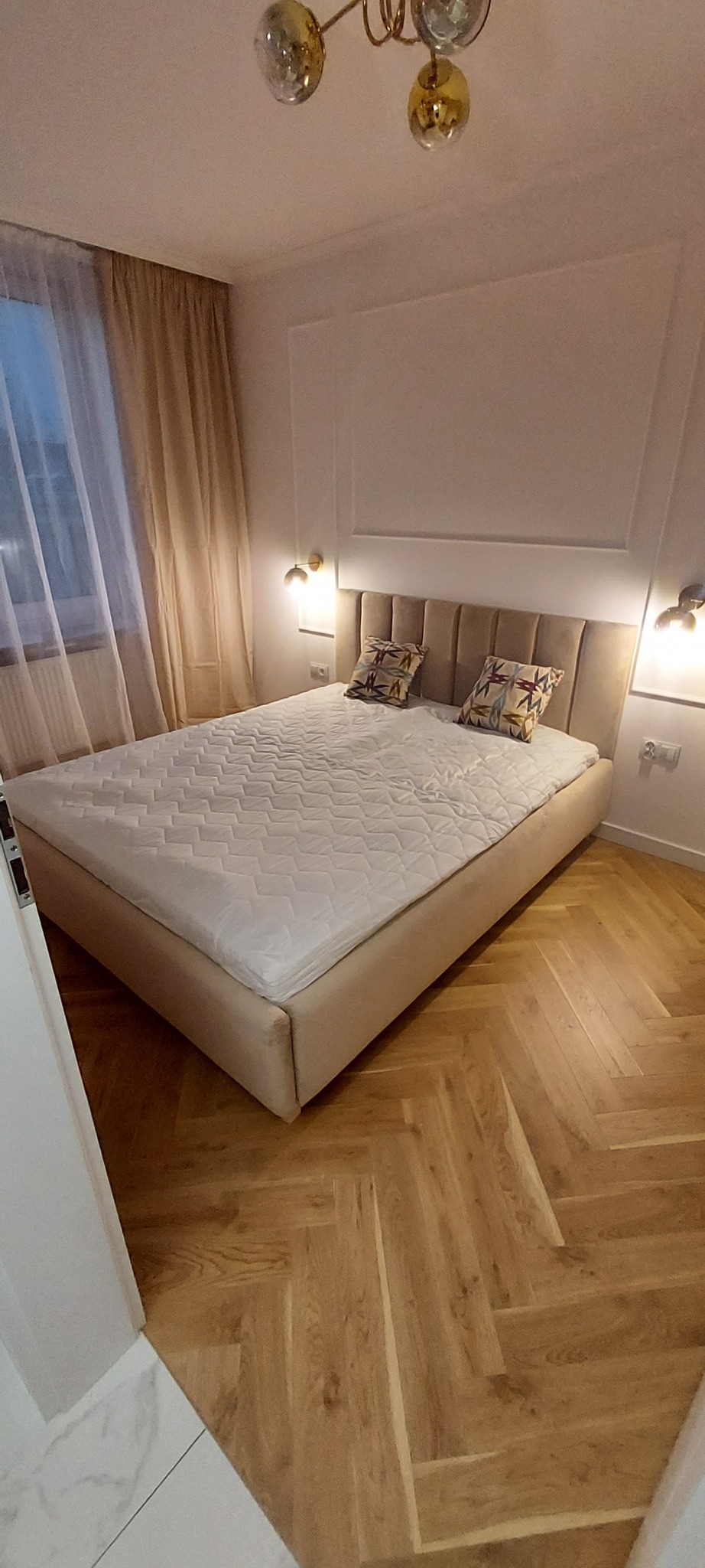 Sypialnia z łóżkiem kontynentalnym z beżowym zagłówkiem, dwoma poduszkami wzorzystymi, parkietem ułożonym w jodełkę, oświetleniem ściennym i zasłonami w odcieniach beżu.