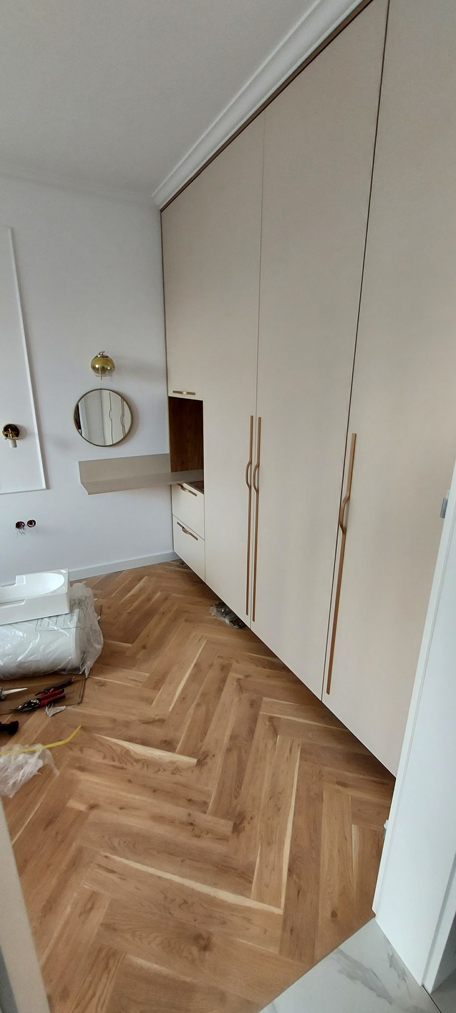 Jasna zabudowa kuchenna z szafkami sięgającymi sufitu, drewnianymi uchwytami i otwartą półką, uzupełniona okrągłym lustrem i złotą lampą na ścianie, na podłodze parkiet ułożony w jodełkę.