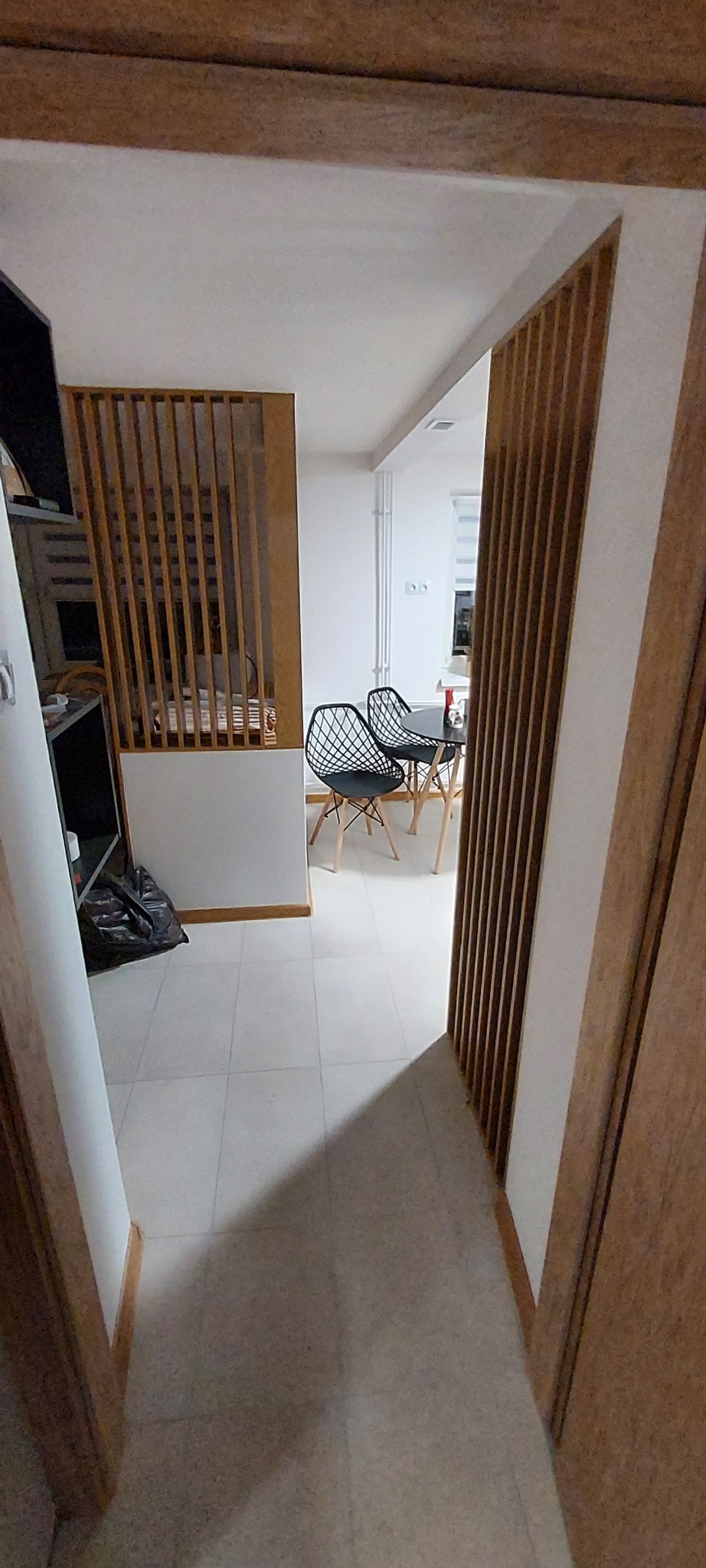 Widok z perspektywy korytarza na wnętrze mieszkania z drewnianymi elementami dekoracyjnymi i ażurowymi ściankami działowymi, oraz jadalnią z czarnymi krzesłami i okrągłym stołem.