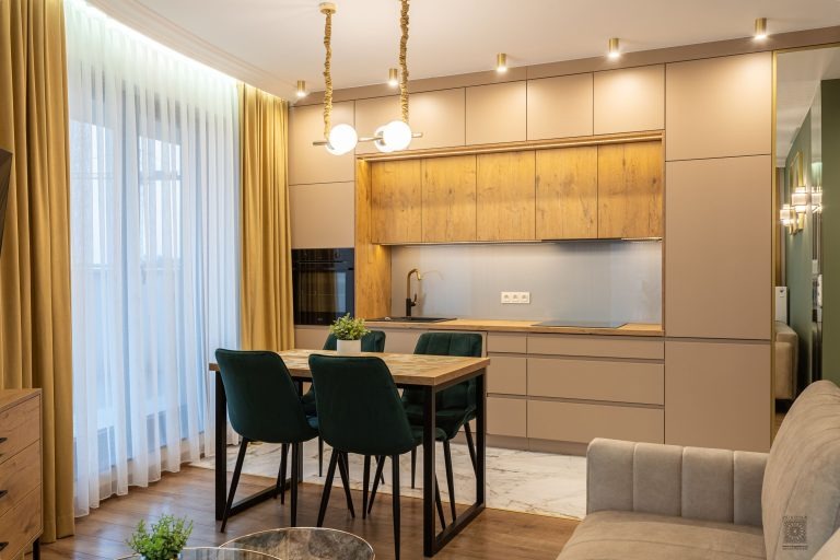 Beżowa kuchnia z drewnianymi akcentami, oświetlona wiszącą lampą z kulistymi kloszami, z ciemnozielonymi krzesłami przy drewnianym stole, widok z salonu.