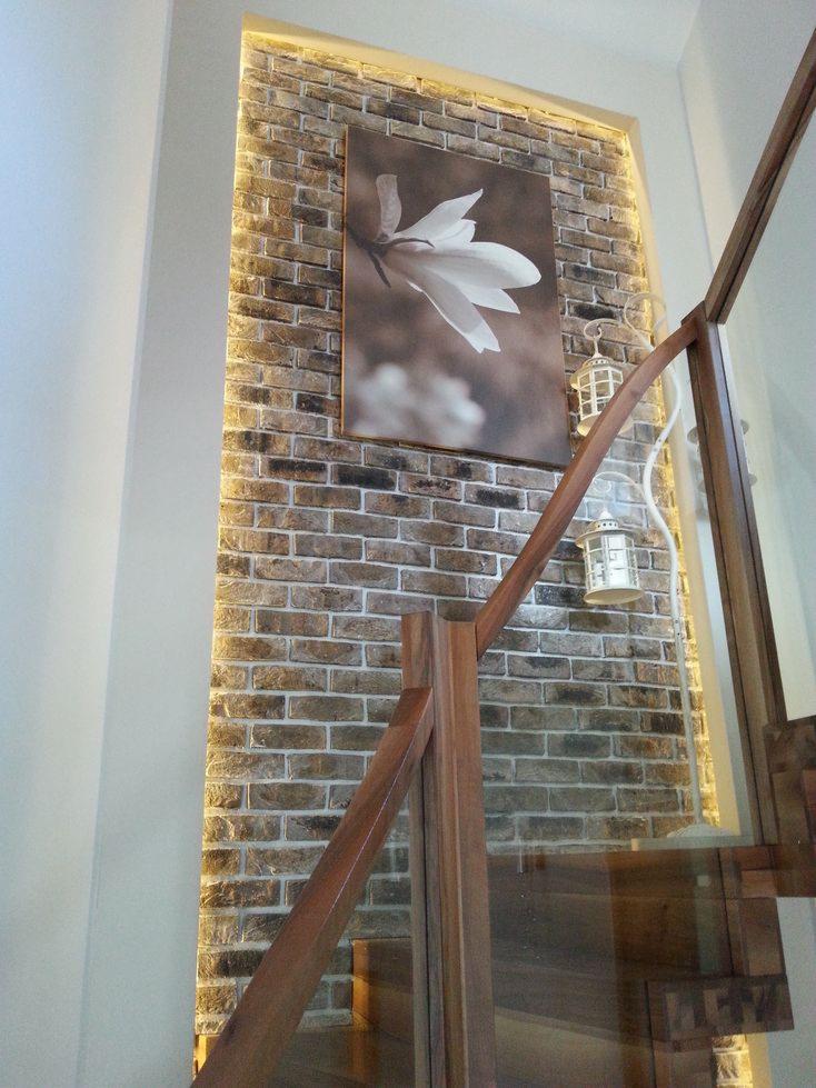 Podświetlana ściana z cegły z obrazem magnolii, drewniane balustrady schodów z elementami szklanymi, dekoracyjne lampiony na ścianie.