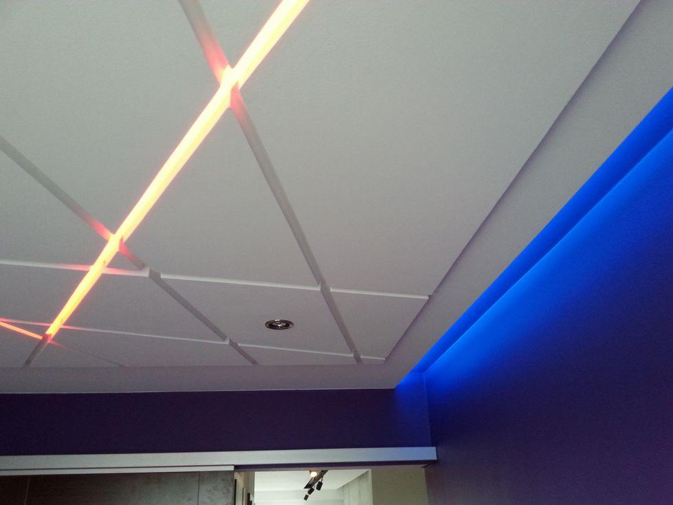 Nowoczesny sufit podwieszany z geometrycznym wzorem, podświetlony liniowym oświetleniem LED w kolorze pomarańczowym i niebieskim, z centralnym punktem świetlnym.