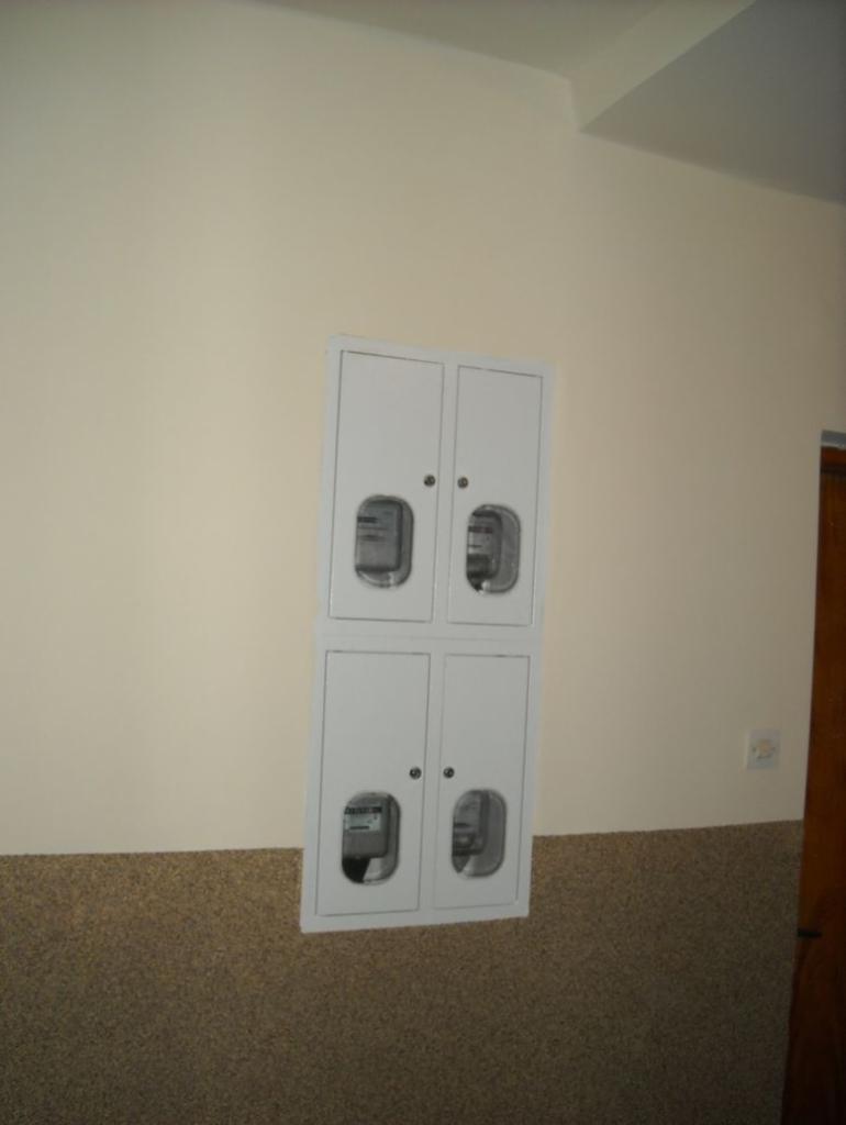 Czterodrzwiowa, biała szafka z licznikami elektrycznymi zamontowana na ścianie z beżową górną i brązową dolną częścią, widoczne liczniki za przeszklonymi drzwiczkami.