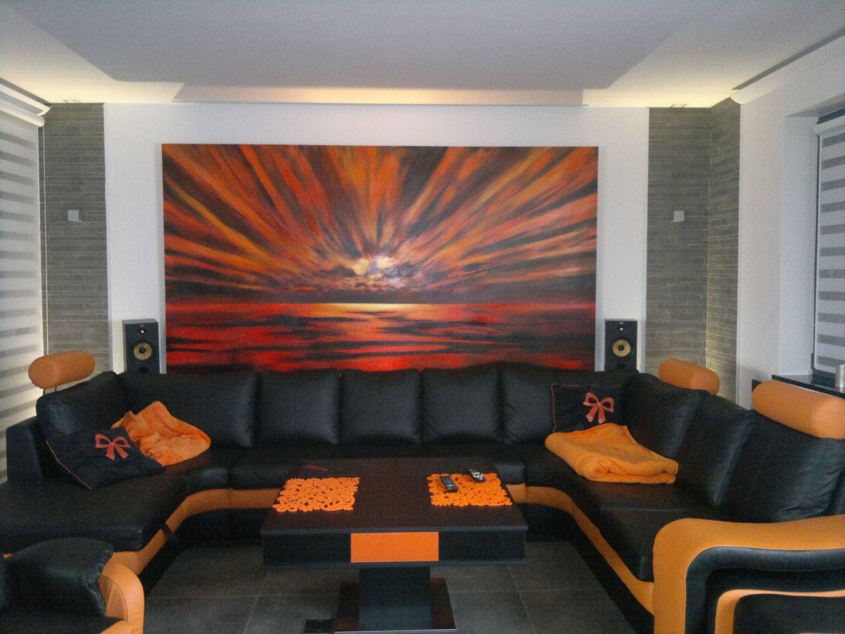 Nowoczesny salon z narożną sofą w kolorze czarnym i pomarańczowym, obraz na ścianie z motywem zachodu słońca, dwa głośniki po bokach obrazu, rolety w oknach.