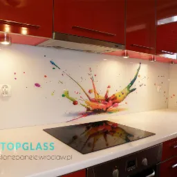 Szklany panel kuchenny z motywem kolorowej eksplozji farb, zamontowany nad płytą grzewczą w nowoczesnej kuchni z czerwonymi szafkami.