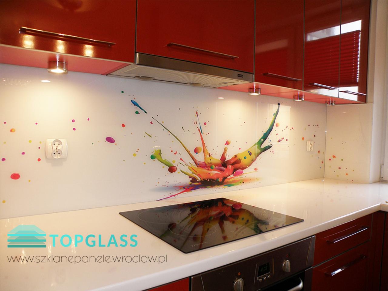 Szklany panel kuchenny z motywem kolorowej eksplozji farb, zamontowany nad płytą grzewczą w nowoczesnej kuchni z czerwonymi szafkami.