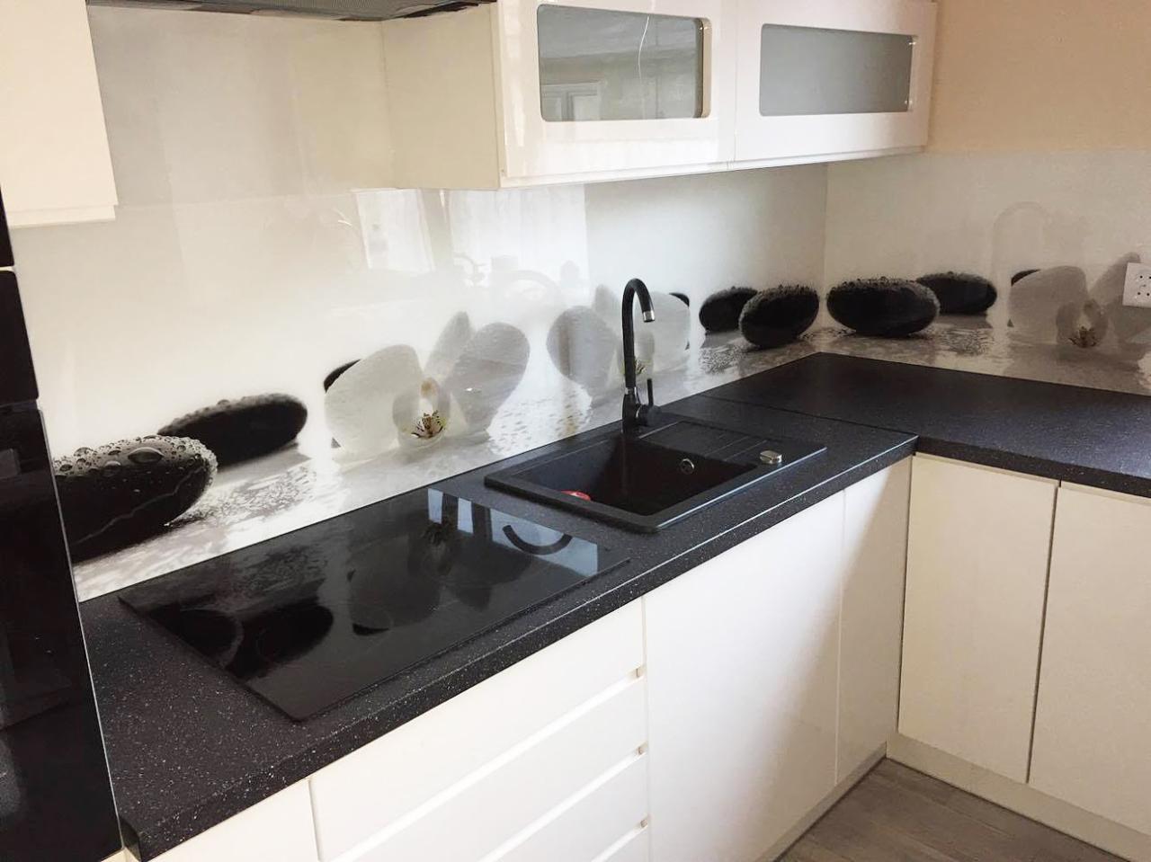 Nowoczesna kuchnia z białymi szafkami, czarnym blatem w drobny brokat i szklanym panelem nad blatem z motywem białych orchidei i czarnych kamieni.