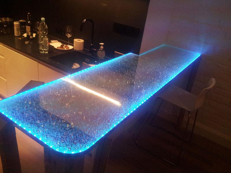 Szklany blat baru podświetlony niebieskimi diodami LED, z widocznym fragmentem blatu kuchennego z umywalką i akcesoriami.