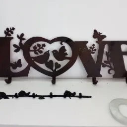 Metalowy wieszak ścienny w kształcie napisu LOVE z motywami ptaków, serca i motyli, wykonany z ciemnego metalu z widoczną fakturą, umieszczony na białym tle.