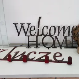 Metalowe dekoracje ścienne: napis 'Welcome Home' w kolorze czarnym, wieszak na klucze z napisem 'Klucze' w kolorze czerwonym oraz ozdobny drewniany element z metalowymi akcentami, ustawione...