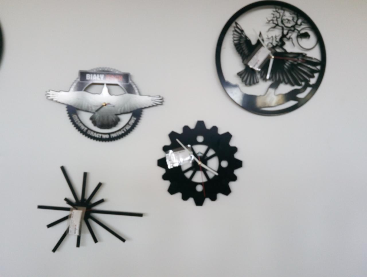 Dekoracyjne, metalowe zegary ścienne o różnych wzorach: orzeł w locie z napisem 'Biały', ptak w okręgu z motywem roślinnym, zębatka oraz abstrakcyjna kompozycja z promieniami.