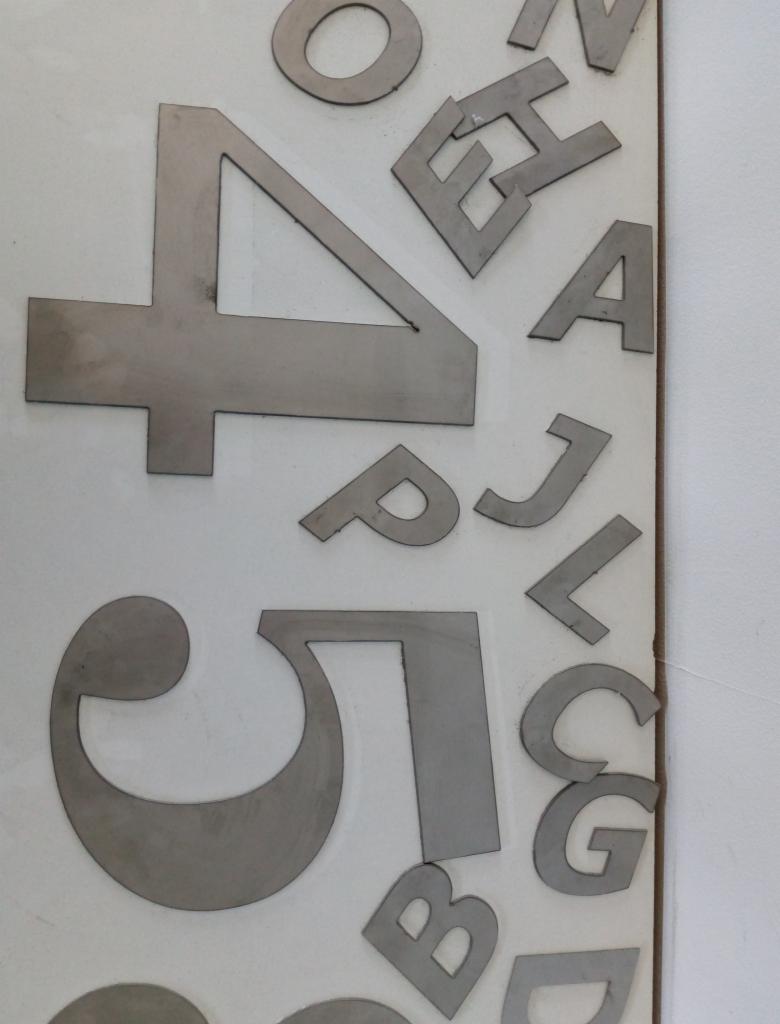 Metalowe litery i cyfry ułożone na białej powierzchni, wykonane przez firmę ze Starego Zamościa. Widoczne kształty: 4, 5, O, H, A, P, J, L, G, B, D oraz inne fragmenty alfabetu.