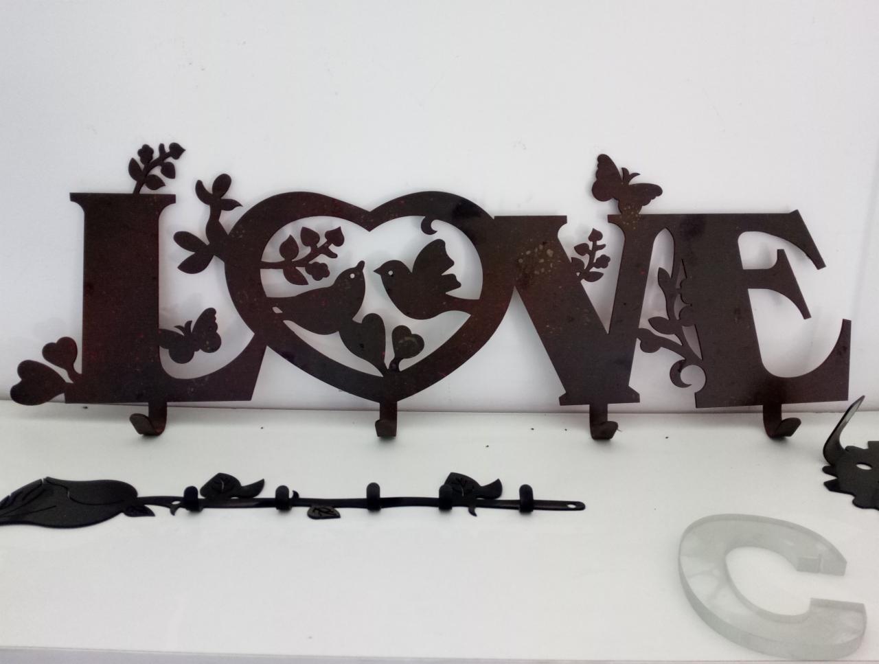 Metalowy wieszak ścienny w kształcie napisu LOVE z motywami ptaków, serca i motyli, wykonany z ciemnego metalu z widoczną fakturą, umieszczony na białym tle.