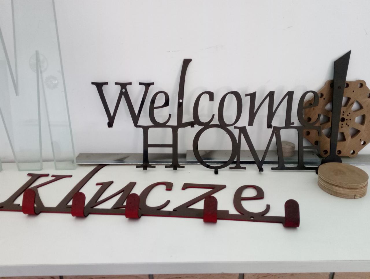 Metalowe dekoracje ścienne: napis 'Welcome Home' w kolorze czarnym, wieszak na klucze z napisem 'Klucze' w kolorze czerwonym oraz ozdobny drewniany element z metalowymi akcentami, ustawione...