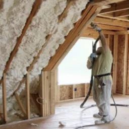Pracownik w kombinezonie natryskuje pianę PUR na poddasze domu w trakcie budowy, widoczne drewniane elementy konstrukcyjne i folia ochronna na podłodze.