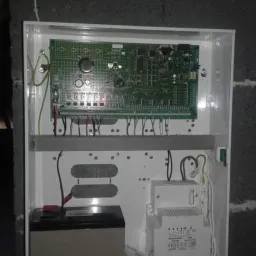 Centrala alarmowa z akumulatorem i transformatorem umieszczona w białej obudowie na szarej ścianie z cegieł.