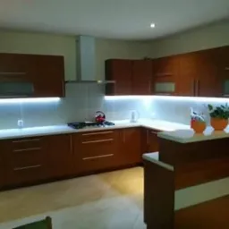 Kuchnia z drewnianymi szafkami i białym blatem, podświetlona listwami LED pod szafkami wiszącymi, z roślinami doniczkowymi na blacie.