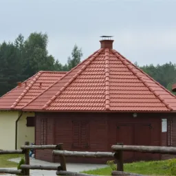 Czerwona dachówka ceramiczna na drewnianym budynku z kominem, widoczne ogrodzenie i las w tle, pochmurny dzień.