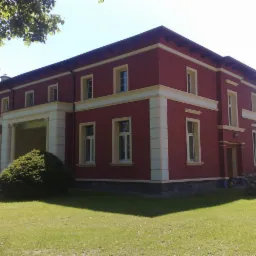 Dwukondygnacyjny budynek z czerwonymi ścianami i kremowymi elementami dekoracyjnymi, widziany z perspektywy trawnika w słoneczny dzień. Widoczny fragment opony w lewym dolnym rogu.