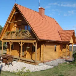 Jasny, dwukondygnacyjny dom z drewna z czerwonym dachem, balkonem z kwiatami i werandą, otoczony zielenią i małą architekturą ogrodową.