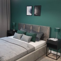 DeKa Denys Peresada - Nowoczesna sypialnia z łóżkiem z szarą tapicerką, zieloną ścianą i minimalistycznymi grafikami w czarnych ramkach, lampka nocna z abażurem w kolorze ściany.