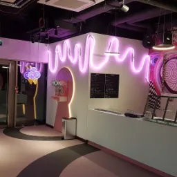 Nowoczesne wnętrze kawiarni z neonowymi dekoracjami, menu na ścianie i ladą. Widoczne elementy wystroju w odcieniach różu i czerni.