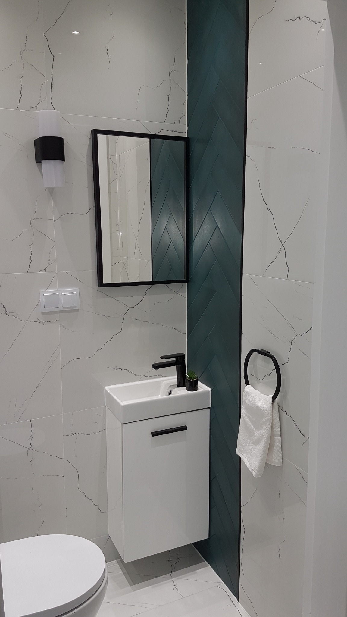 Nowoczesna łazienka z białą umywalką nablatową, czarną armaturą, lustrem w czarnej ramie i akcentem ściany z ciemnozielonej glazury ułożonej w jodełkę.