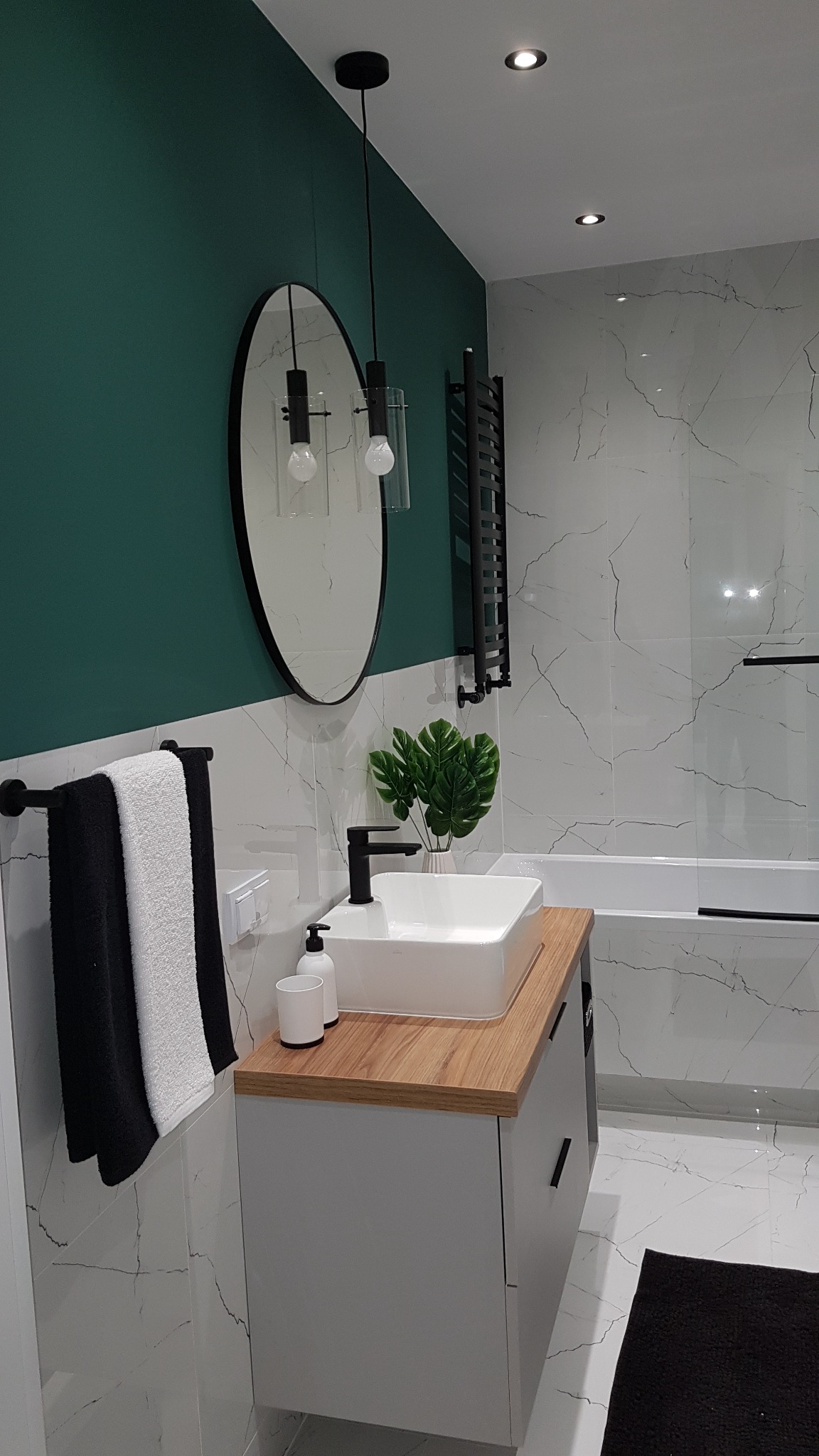 Nowoczesna łazienka z białymi marmurowymi płytkami, zieloną ścianą, owalnym lustrem, wiszącą lampą, białą umywalką nablatową na drewnianym blacie i czarnymi akcentami.