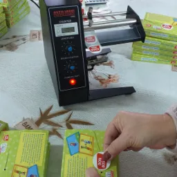 Naklejanie etykiety 'Don Bosco' na zielone pudełko z kartami edukacyjnymi przy użyciu automatycznego dyspensera etykiet z wyświetlaczem LCD pokazującym licznik 2042.