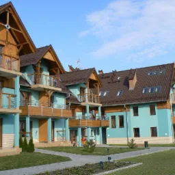 Kompleks apartamentów wakacyjnych z balkonami, turkusową elewacją i drewnianymi elementami wykończeniowymi, widok z zewnątrz na zadbany trawnik i chodnik.