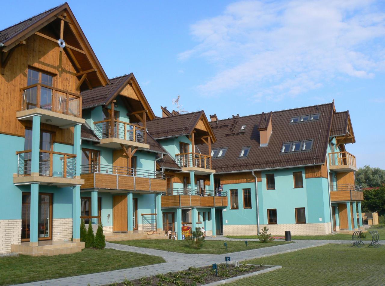 Kompleks apartamentów wakacyjnych z balkonami, turkusową elewacją i drewnianymi elementami wykończeniowymi, widok z zewnątrz na zadbany trawnik i chodnik.