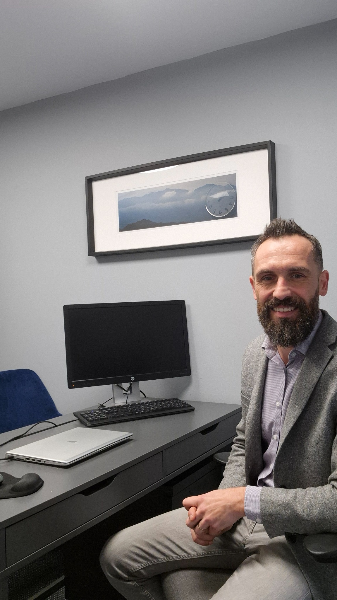 Uśmiechnięty mężczyzna z brodą siedzi przy biurku z komputerem, laptopem i klawiaturą w biurze o szarych ścianach, na których wisi obraz z górami i zegarem.