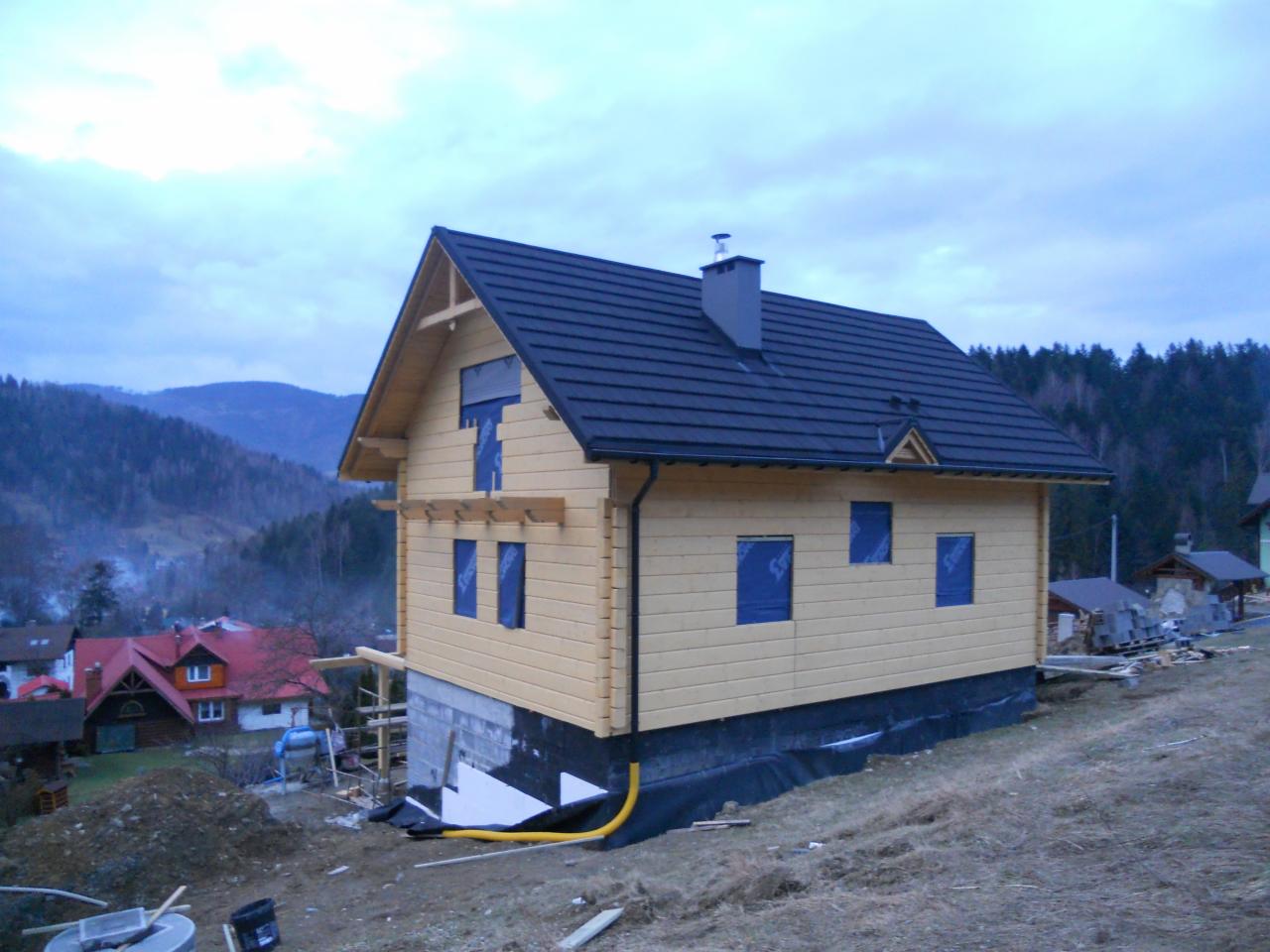 Surowy dom szkieletowy w Szczyrku, z ciemnym dachem i oknami zabezpieczonymi folią, na nierównym terenie w otoczeniu górskiego krajobrazu.