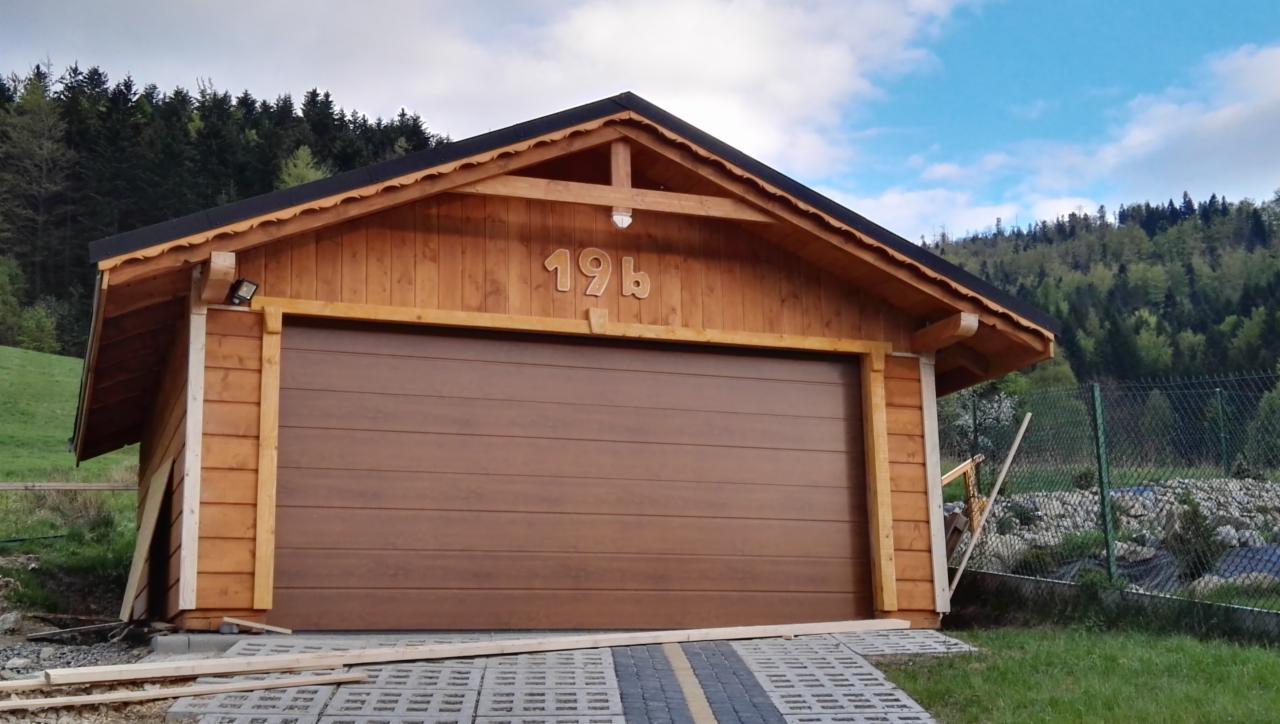 Drewniany garaż z brązową bramą segmentową, numer 19b nad wejściem, na tle zalesionego wzgórza i błękitnego nieba.