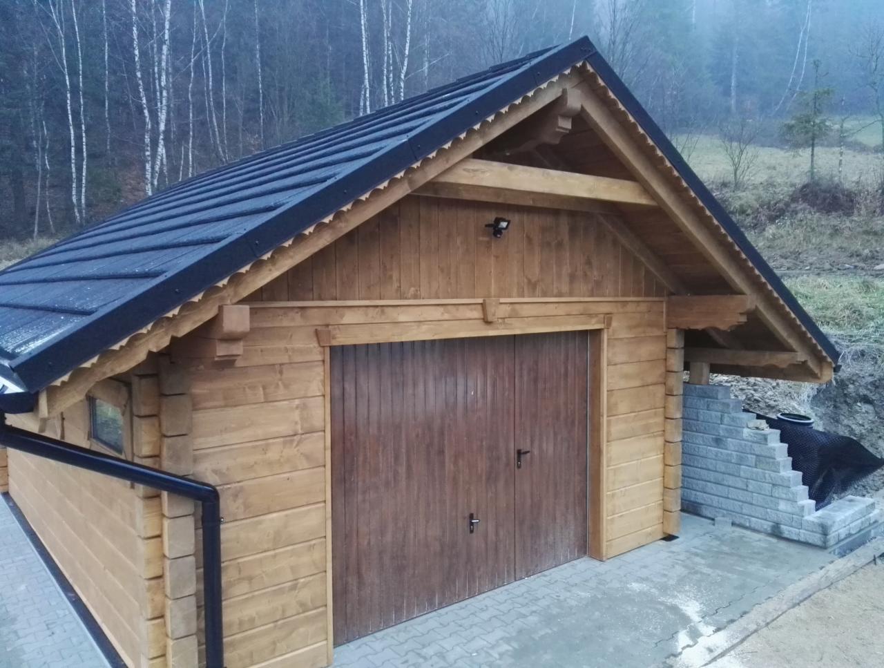 Drewniany budynek gospodarczy z dwuspadowym dachem pokrytym czarną blachodachówką, widoczne detale wykończeniowe dachu i elewacji, w tle zalesione wzgórze we mgle.