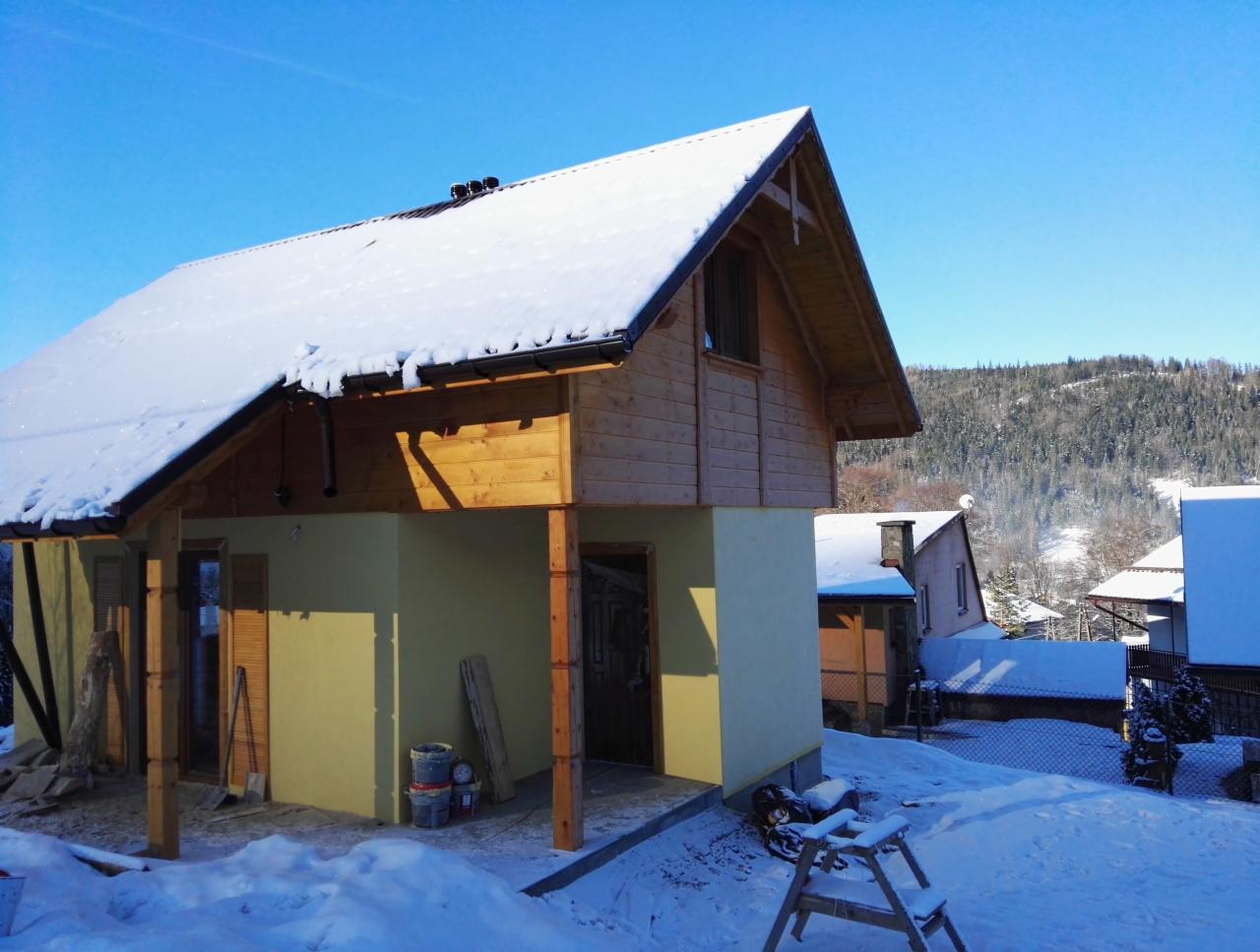 Surowy stan domu z drewnianą konstrukcją szkieletową w zimowej scenerii, częściowo otynkowany, widoczne rusztowania i śnieg na dachu.