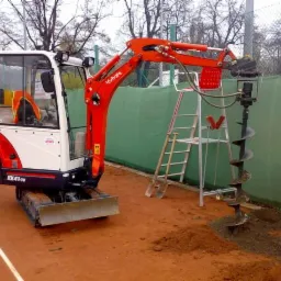 Mała koparka Kubota z wiertnicą hydrauliczną wykonuje odwiert w ziemi na korcie tenisowym, obok stoi drabina aluminiowa.