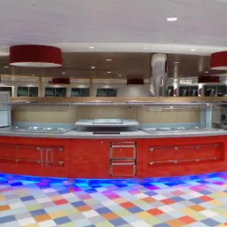 Okrągły bar samoobsługowy w kolorze czerwonym z niebieskim oświetleniem LED u dołu, widoczne urządzenia do napojów i podgrzewacze potraw, podłoga w kolorowe kwadraty.