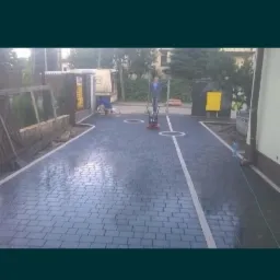 Układanie ciemnoszarej kostki brukowej na podjeździe, widoczny pracownik z zagęszczarką w trakcie pracy, obok białe linie oddzielające miejsca parkingowe.